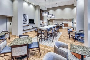 Um hall moderno de hotel com uma longa mesa de refeições compartilhada, vários reservados para refeições e uma área de recepção.