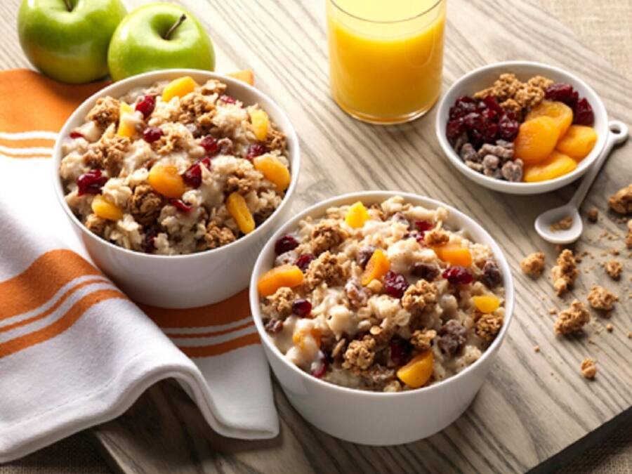 Two bowls of oatmeal with fruits and granola, a glass of orange juice, and two green apples on a wooden table.