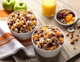 Dos tazones de avena con frutas y granola, un vaso de zumo de naranja y dos manzanas verdes sobre una mesa de madera.