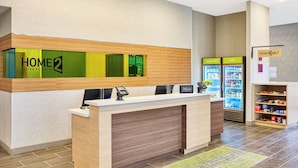 A hotel reception area with a reception desk, a green 'HOME2' sign, a minibar, and a self-service beverage machine.