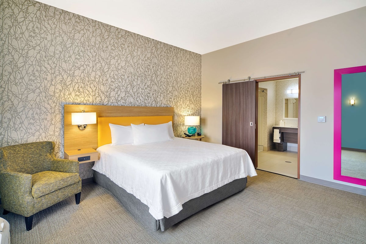 Une chambre d’hôtel avec un grand lit, une chaise, une table de chevet avec des lampes, une salle de bain visible à travers une porte ouverte et un mur à motifs.