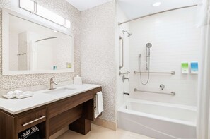 Une salle de bain moderne avec un lavabo blanc, un miroir et une baignoire. On y trouve également des barres d'appui et une pomme de douche.