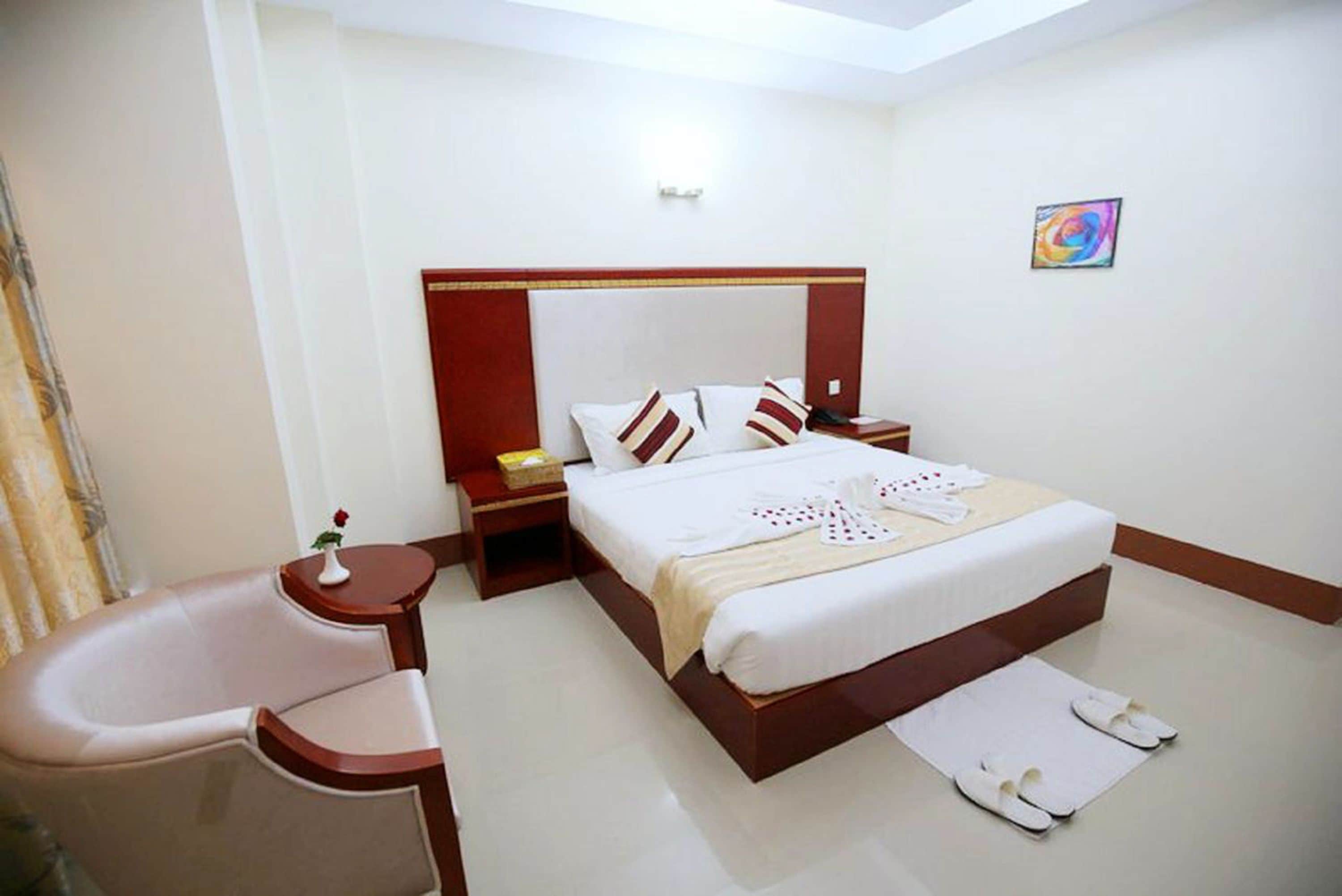 A hotel room with a bed, a bedside table, a chair, and a small table with a vase.