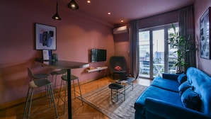 Een moderne woonkamer met een blauwe bank, een eettafel met stoelen, een televisie en een groot raam met uitzicht op de stad.