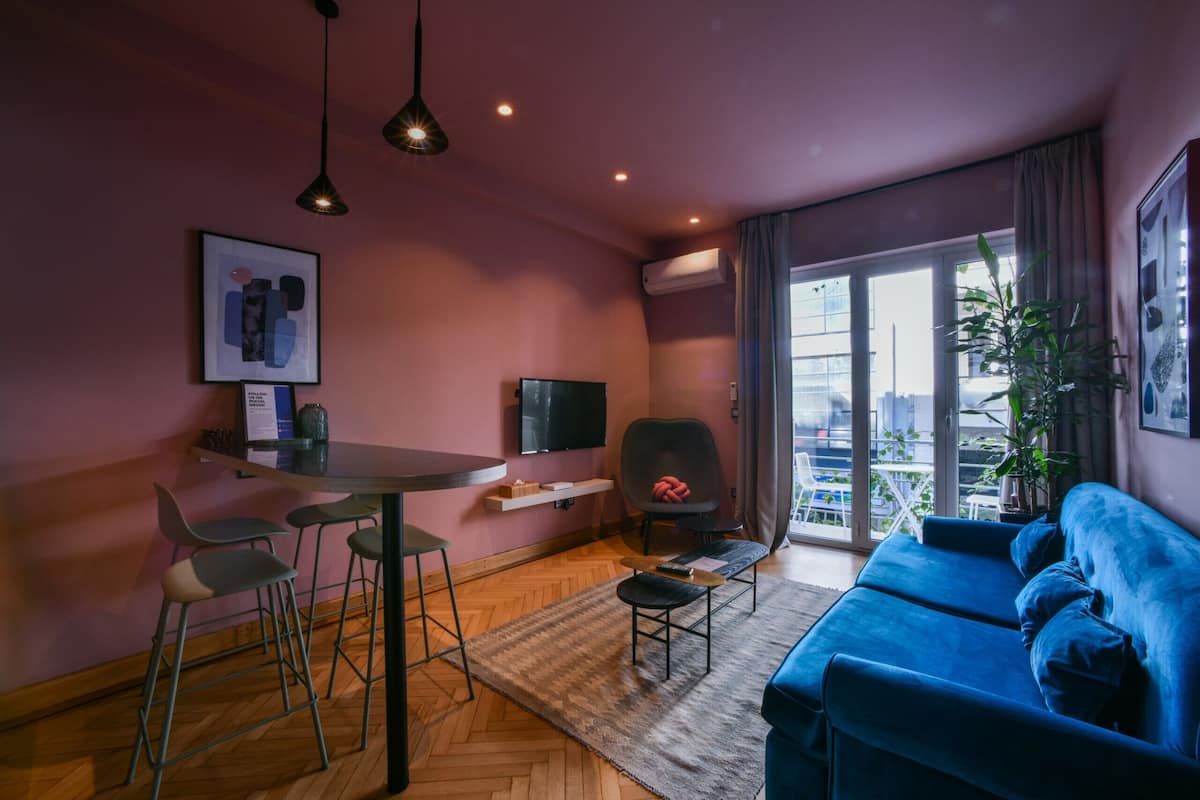 Een moderne woonkamer met een blauwe bank, een eettafel met stoelen, een televisie en een groot raam met uitzicht op de stad.