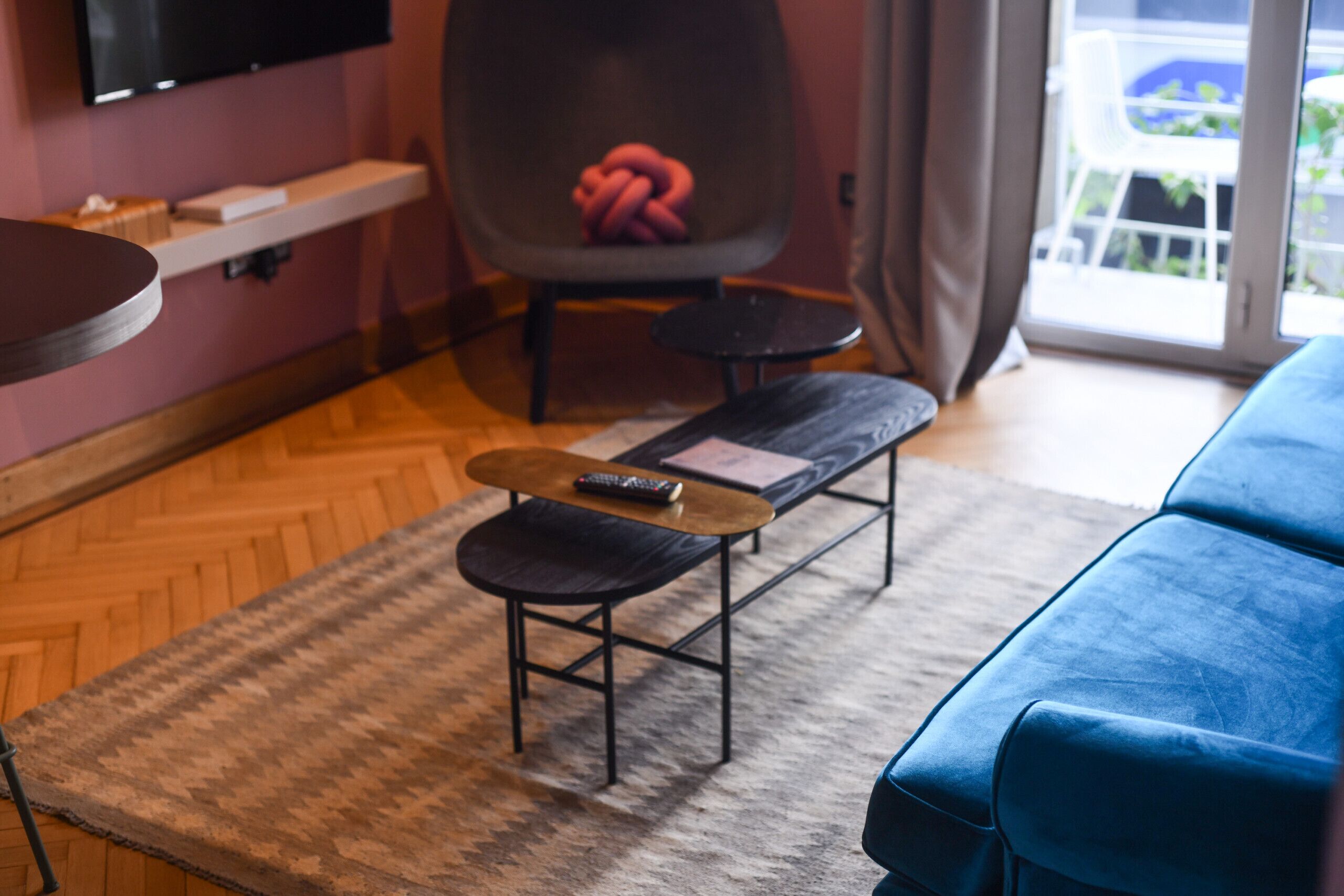 Un salon moderne avec un canapé bleu, une table basse en bois sur laquelle repose une télécommande, un tapis à motifs, un fauteuil gris agrémenté d’un objet rose, et un téléviseur fixé au mur.