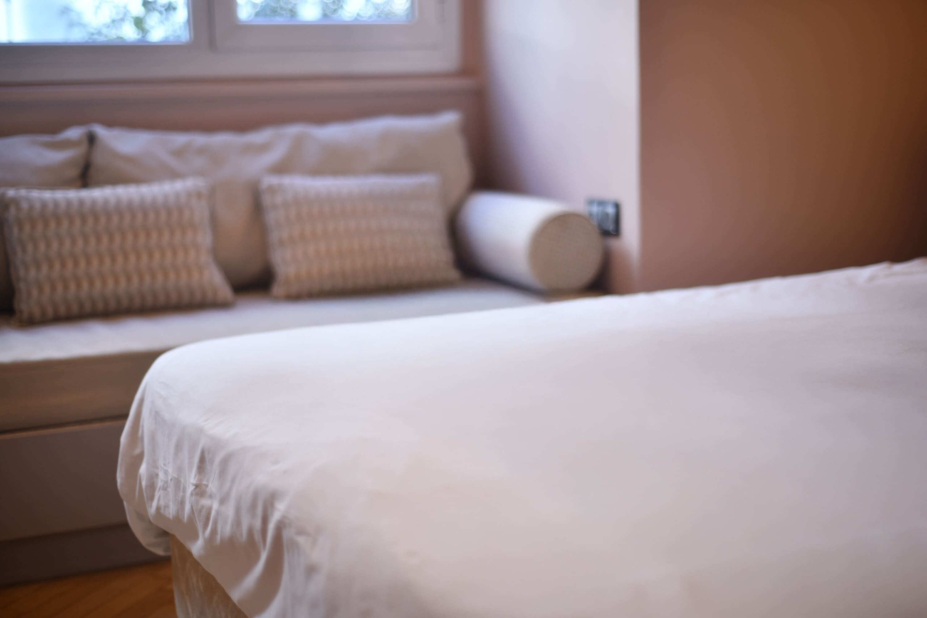 A bed with white bedding in a room with a window and a sofa with cushions.