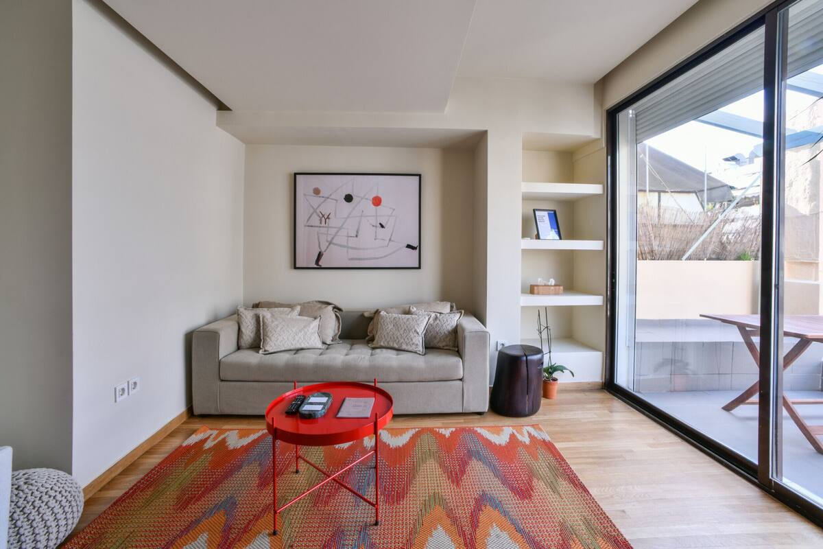 Een moderne woonkamer met een bank, een rode bijzettafel, een groot abstract schilderij en een glazen schuifdeur naar een buitenruimte.