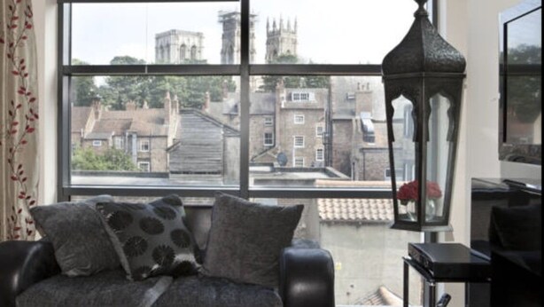A room with a large window, a sofa with patterned cushions, a table with a remote control, and a decorative lantern. - Churchill Apartments (York)