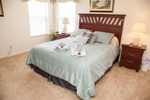 A neatly made bed with a quilt, pillows, and folded towels. There is a nightstand with a lamp and a small drawer. A window with blinds is visible, and a plant is placed beside the window. - Ly53791 - Emerald Island - 6 Bed 5 Baths Villa (Kissimmee)