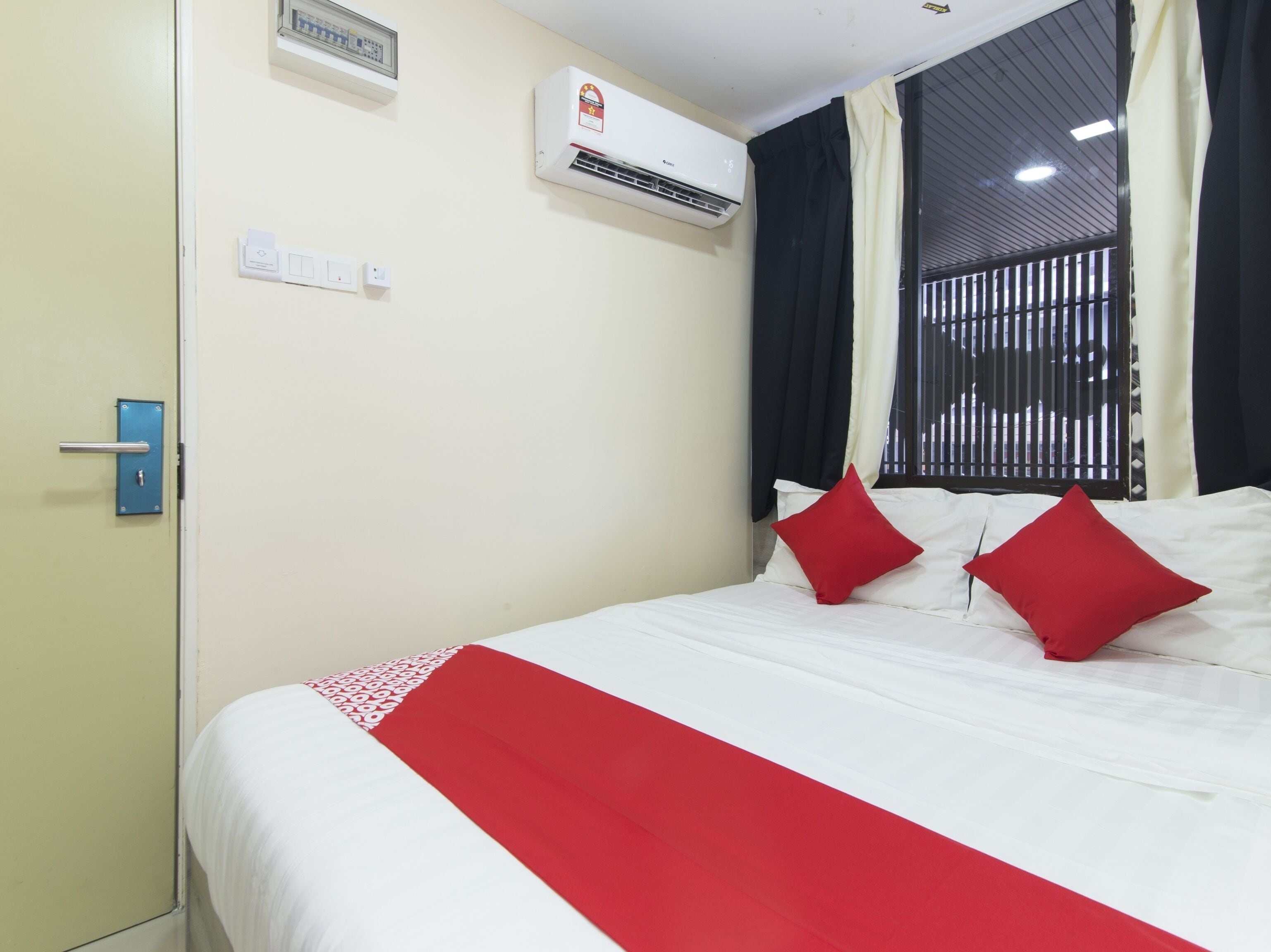 A hotel room with a bed, a wall-mounted air conditioner, a window with black curtains, and a door with a blue handle.