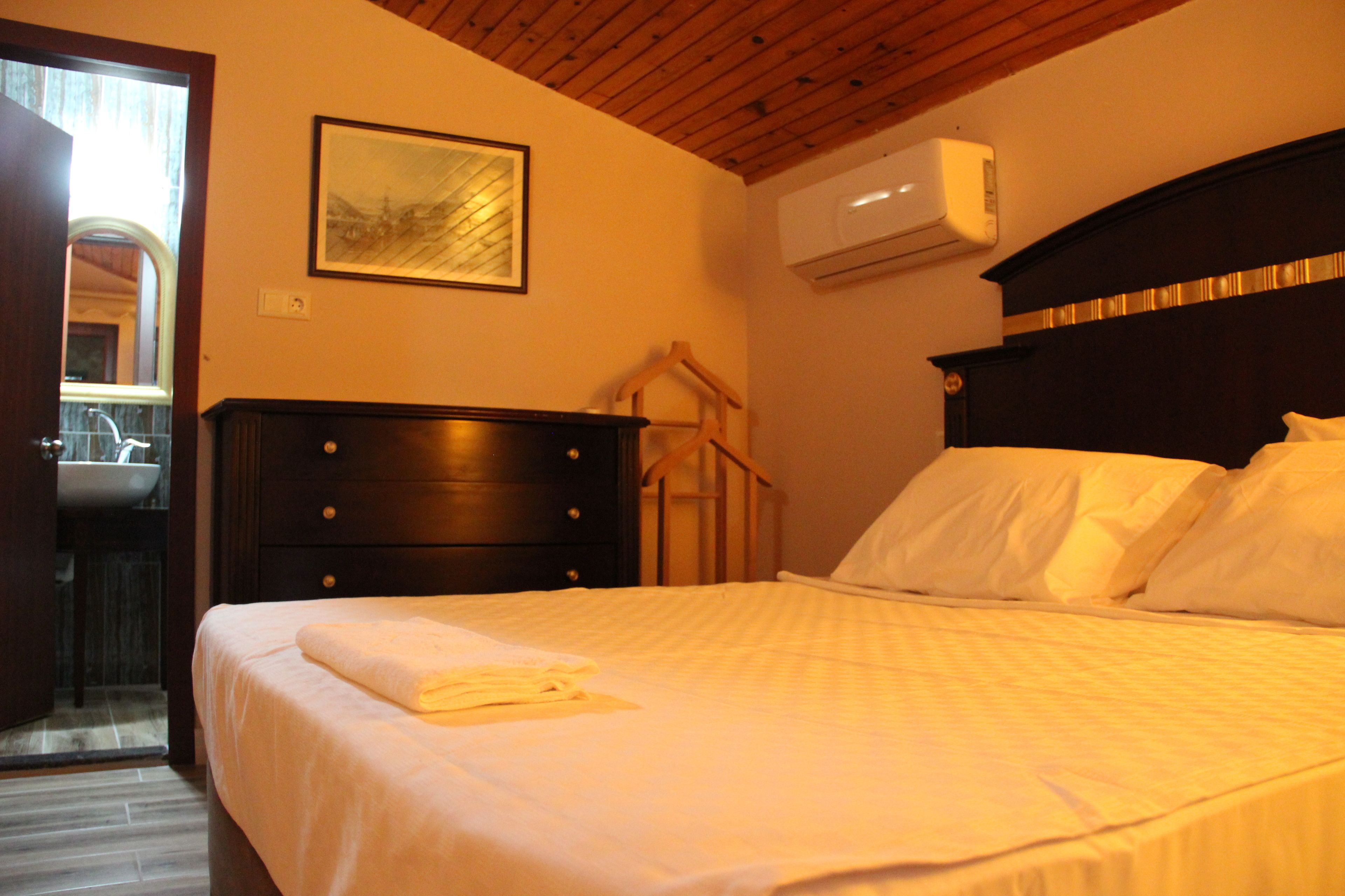 A neatly made bed with a white comforter and a folded towel, a dark wooden dresser, a framed picture on the wall, and an air conditioning unit mounted above the bed.
