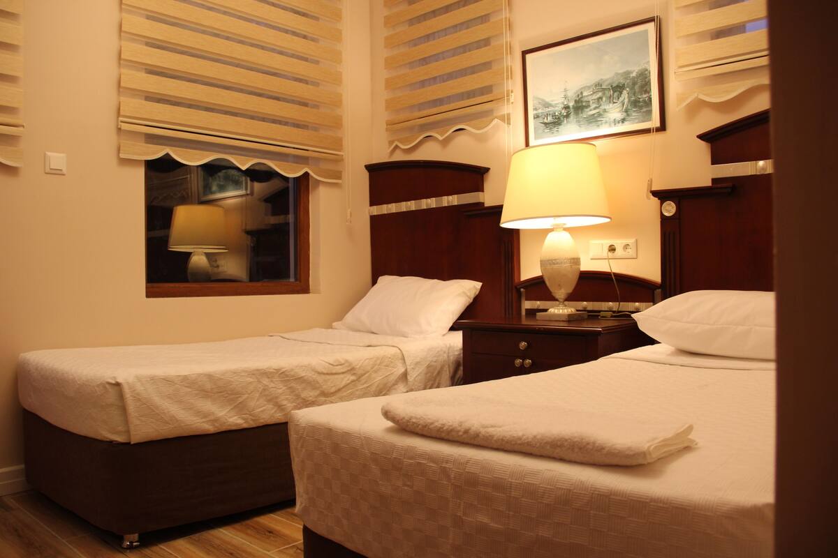 Una camera d'albergo con due letti singoli, un comodino con lampada, una finestra con persiane e un quadro appeso al muro.