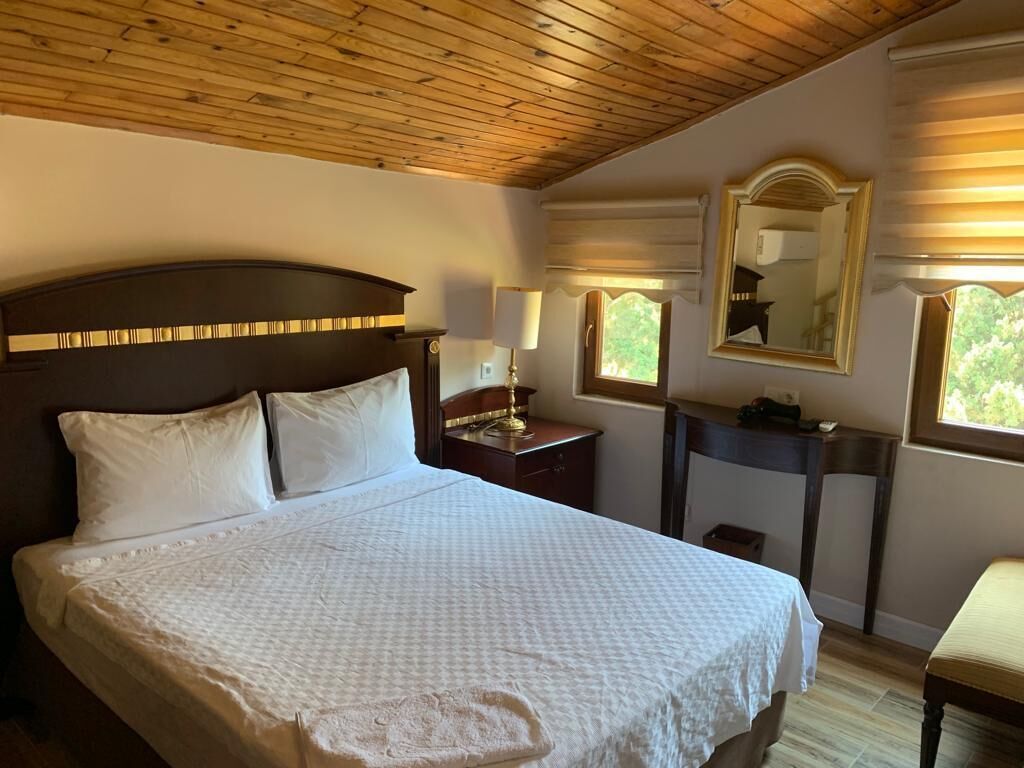 Une chambre à coucher avec un grand lit, un plafond en bois, une table de chevet avec une lampe, un miroir et une fenêtre avec des stores.