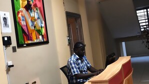 A man sitting at a desk in an office with a digital clock displaying 8:41:00. - JLW Hostel (Berekuso)