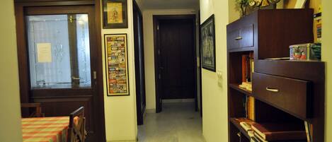 A hallway with a wooden door, a framed poster on the wall, and a bookshelf with books and a canister.