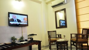 A hotel room with a television mounted on the wall, a wooden bench, two wooden chairs, a small round table, and an air conditioning unit. - OYO Premium Saket Metro Station (New Delhi)