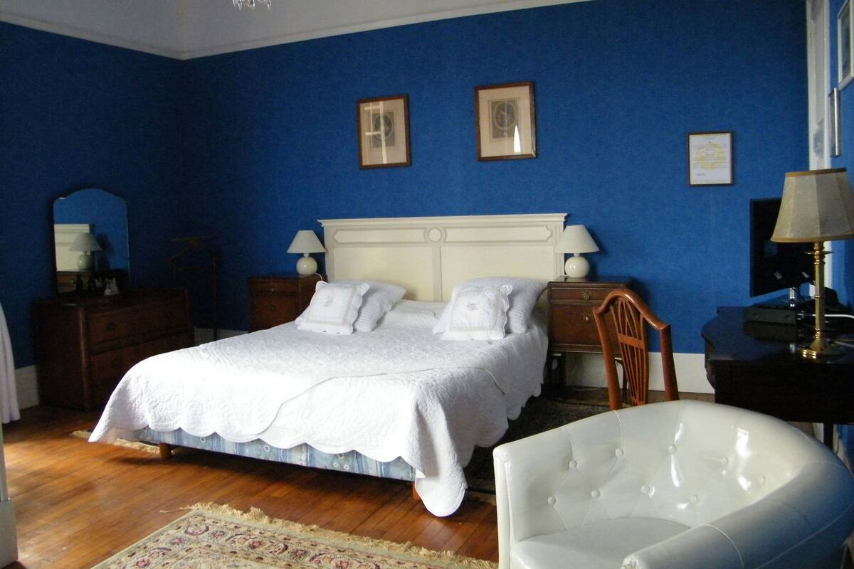 Een slaapkamer met een groot bed, twee nachtkastjes met lampen, een houten stoel en een kroonluchter.