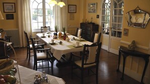 A dining room with a long table set for a meal, surrounded by chairs. There are two windows with curtains, a radiator, and a cabinet with dishes. - Villa des Iris (Saint-Felix)