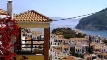 Une ville côtière aux bâtiments aux toits de tuiles rouges, avec un imposant bâtiment jaune doté d’un balcon, et offrant une vue sur la mer et les collines.