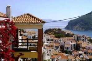 A coastal town with terracotta-roofed buildings, a prominent yellow building with a balcony, and a view of the sea and hills. - Aperanto Galazio (Skopelos)