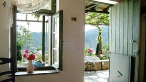 A room with a window overlooking a garden and mountains. There is a chair and a table with a vase of flowers. - Aperanto Galazio (Skopelos)