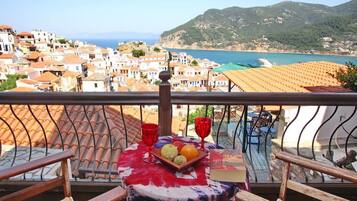 Un balcon avec une table dressée pour deux personnes, donnant sur une ville côtière aux toits de tuiles rouges et offrant une vue sur la mer et les collines.