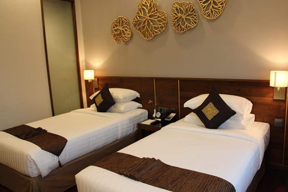 Ein Hotelzimmer mit zwei Betten, jeweils mit weißer Bettwäsche und braunen Überwürfen. Darüber befinden sich dekorative Wandverkleidungen und auf jeder Seite Nachttischlampen.