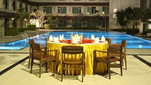 Un área de comedor al aire libre con piscina, mesas dispuestas con manteles blancos y sillas marrones, y un edificio con múltiples ventanas al fondo.