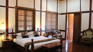 Un dormitorio tradicional con una cama grande, cabecero de madera y un banco al pie de la cama. Se observa un mesita de noche con una lámpara y un teléfono. También se ve una silla de madera y un escritorio con silla.