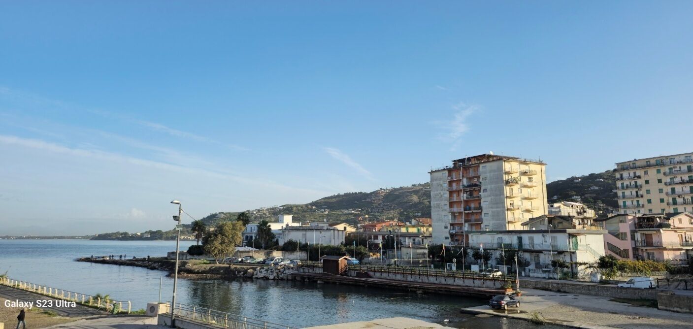 Un paysage urbain côtier avec un bâtiment imposant, une promenade en bord de mer et une chaîne de collines en arrière-plan.