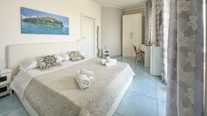 A hotel room with a bed, a desk, a wardrobe, and a painting of a coastal scene. - 100 Passi Dal Mare (Agropoli)