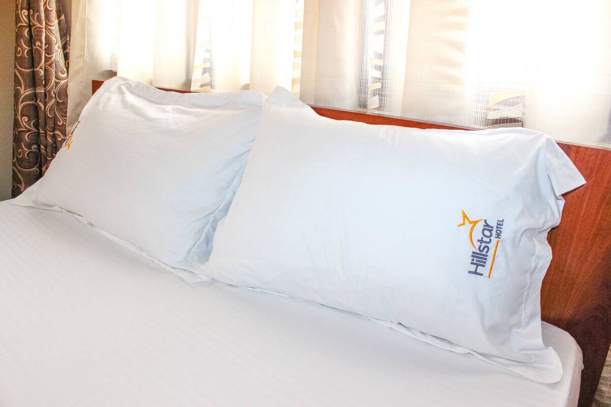 Ein ordentlich bezogenes Bett mit weißen Leinen und einem Kopfkissen mit dem Logo des Hillstar Hotels.