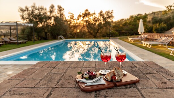 Mesa junto a la piscina con comida y bebidas, rodeada de sillones de descanso y una piscina.