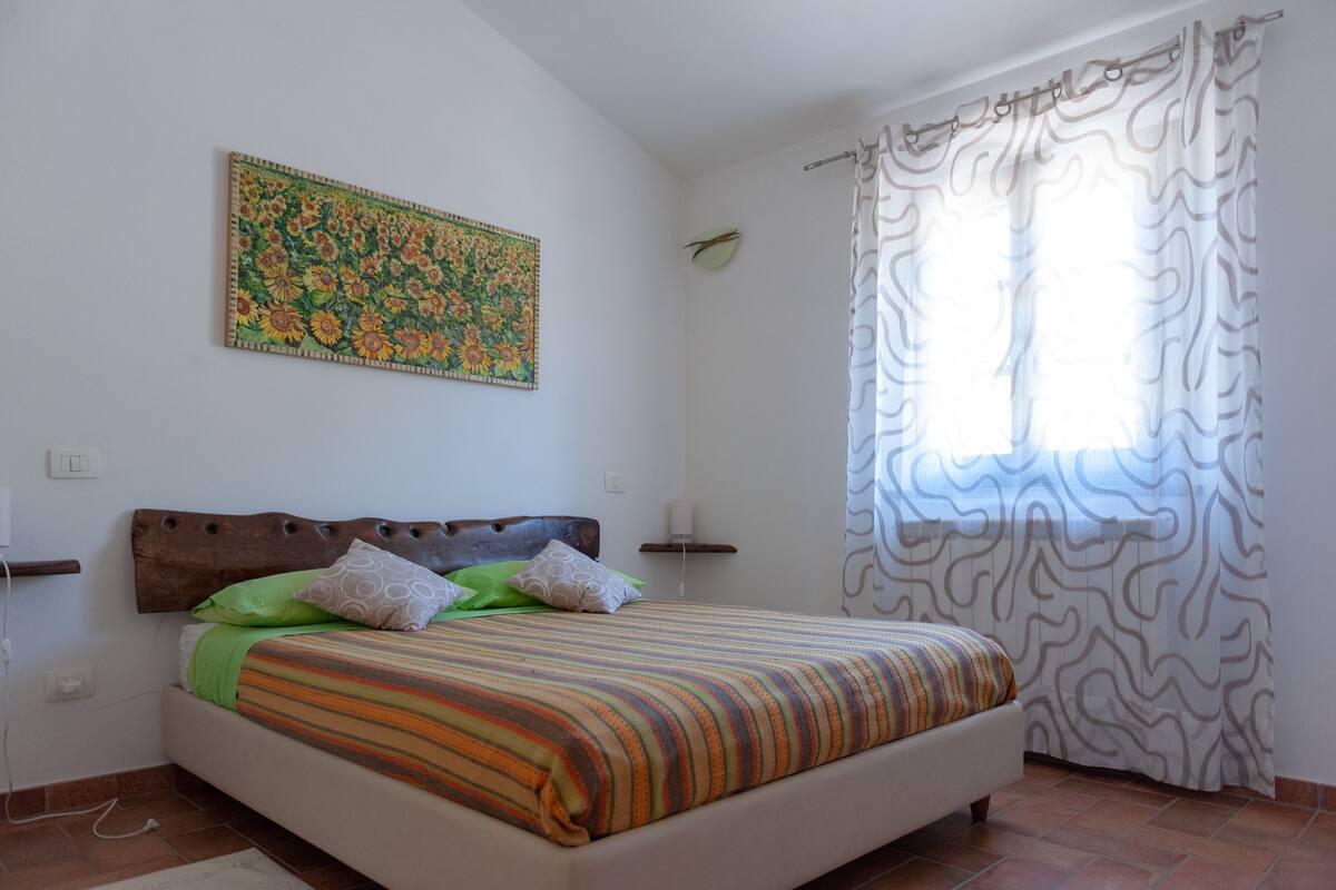 Ein Schlafzimmer mit einem Bett, einem Wandbild, einem Fenster mit gemusterten Vorhängen und einer Steckdose.