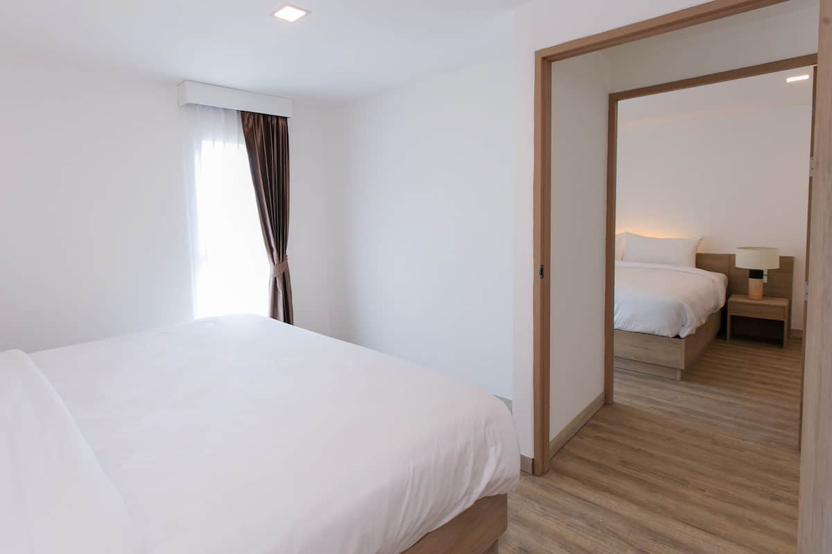 A modern hotel room with a large bed, a wooden floor, and a view through an open door to another room.