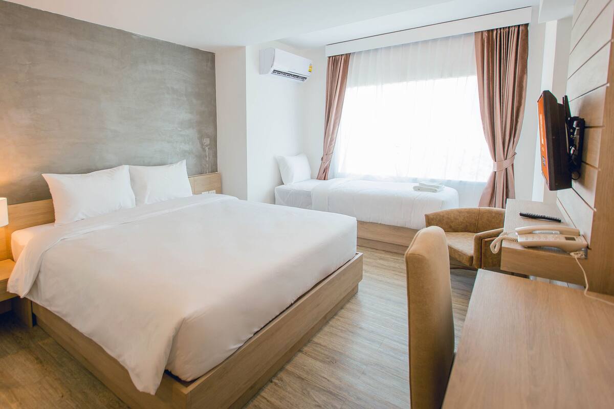 A hotel room with a large bed, a smaller bed, a desk with a telephone, a chair, a nightstand with a lamp, and an air conditioning unit.