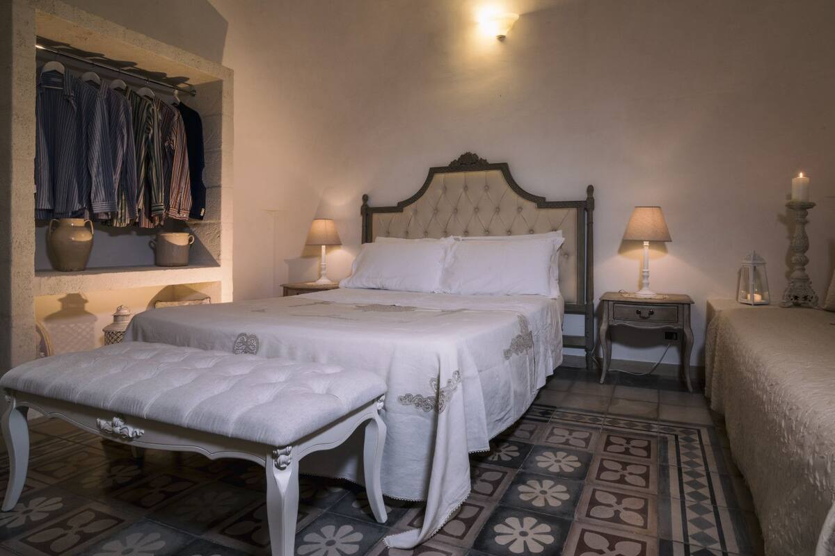 Une chambre à coucher comprenant un lit, deux tables de chevet, une commode, un banc et une étagère avec des vêtements et des objets en céramique.