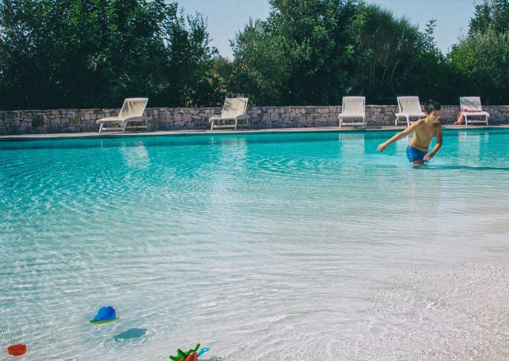 A swimming pool with clear water, surrounded by white lounge chairs and a stone wall.