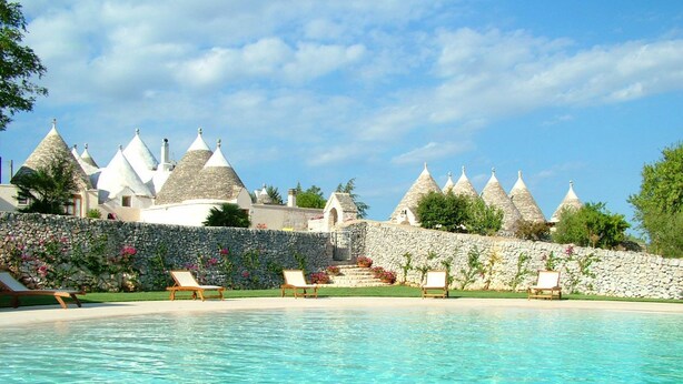 A swimming pool with lounge chairs, a stone wall, and conical-roofed buildings in the background. - Masseria Cappuccini (Ostuni)