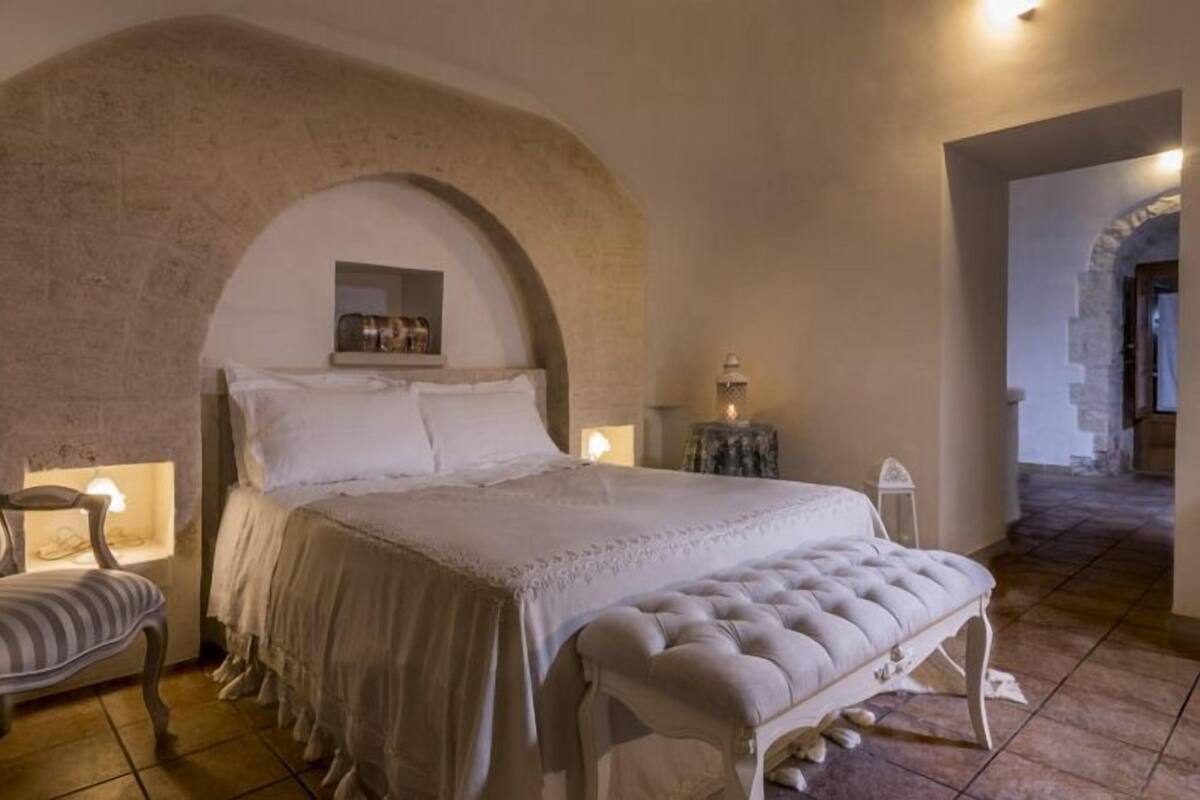 Une chambre avec un grand mur en pierre en arc de cercle, un lit recouvert de draps blancs, un banc et une petite table avec une lampe.