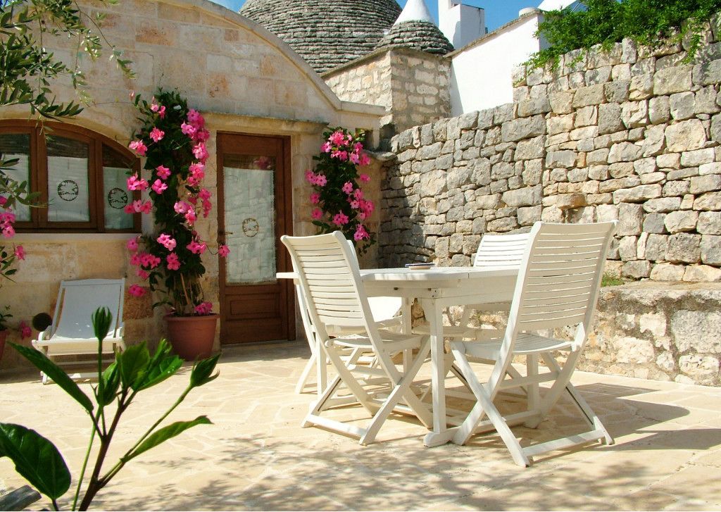 Une terrasse extérieure avec un mur en pierre, une table blanche avec quatre chaises et des plantes en pot ornées de fleurs roses.
