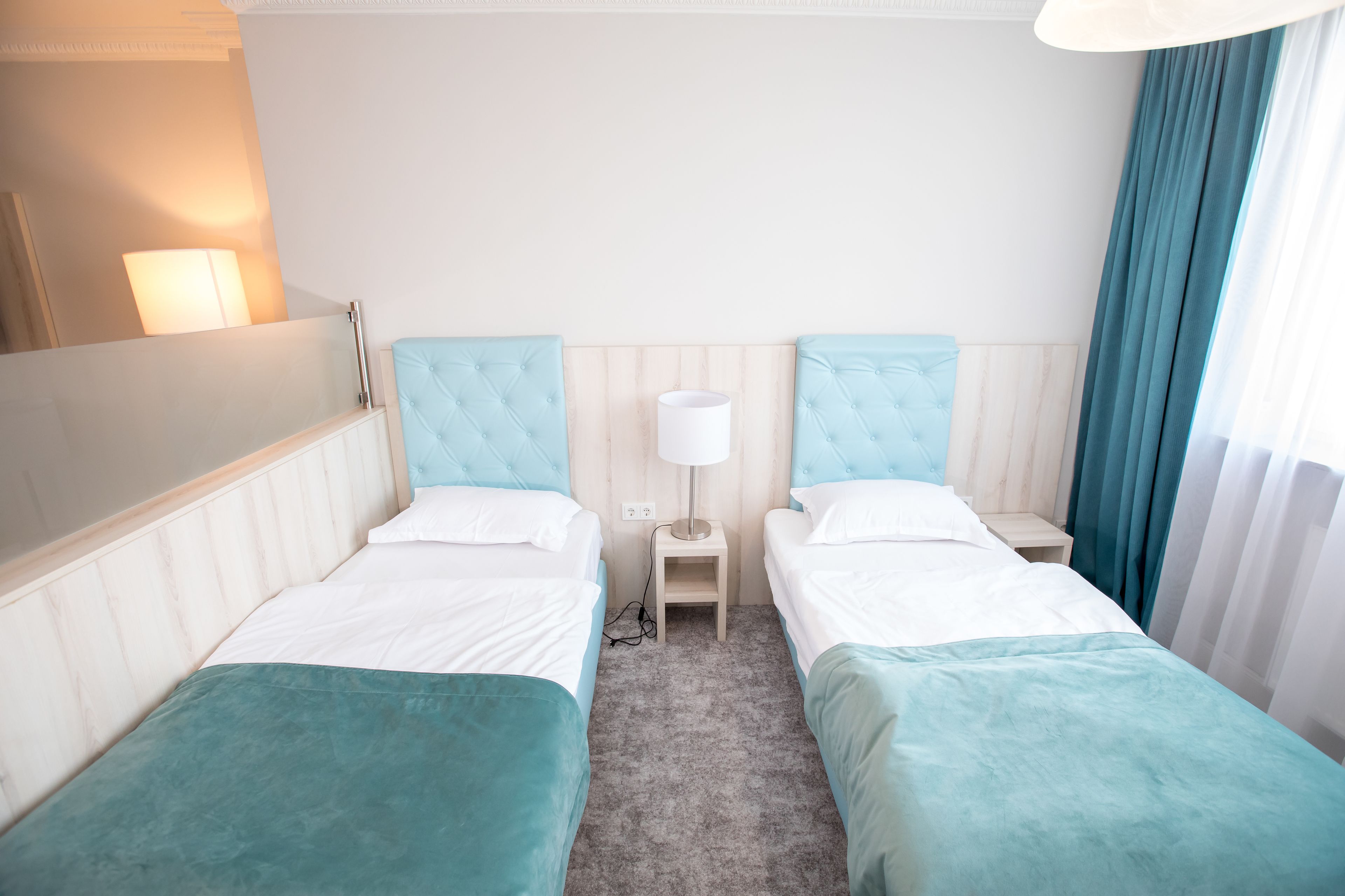 A hotel room with two single beds, a nightstand, a lamp, and a window with curtains.