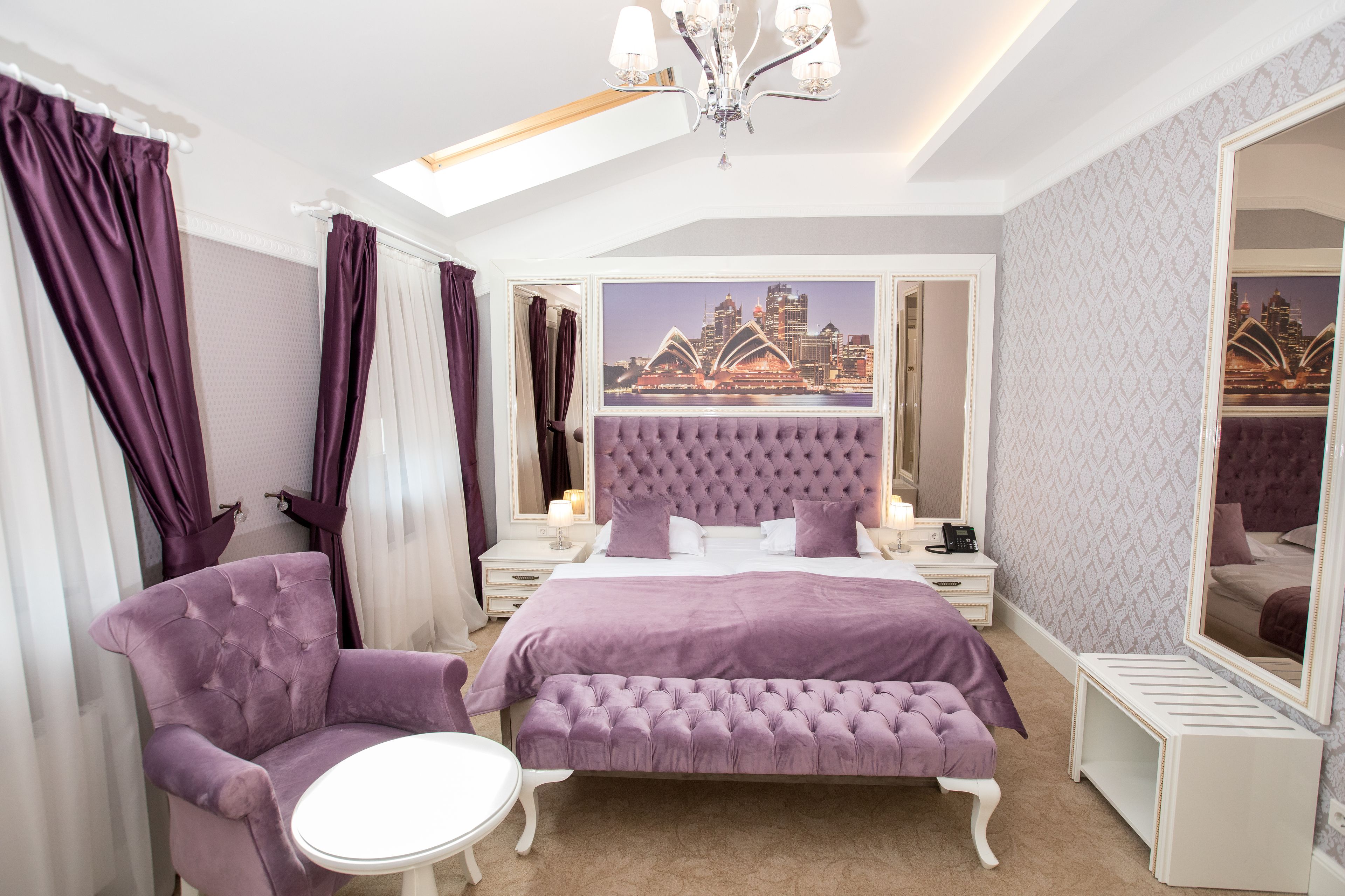 Quarto com cama grande, cabeceira estofada, um banco, uma cadeira, mesa de cabeceira, espelho e vista para a cidade.
