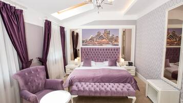 Quarto com cama grande, cabeceira estofada, um banco, uma cadeira, mesa de cabeceira, espelho e vista para a cidade.