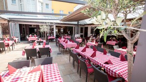 Un espace de restauration en plein air, avec des tables à carreaux et des chaises en osier, situé devant un bâtiment doté de grandes fenêtres.