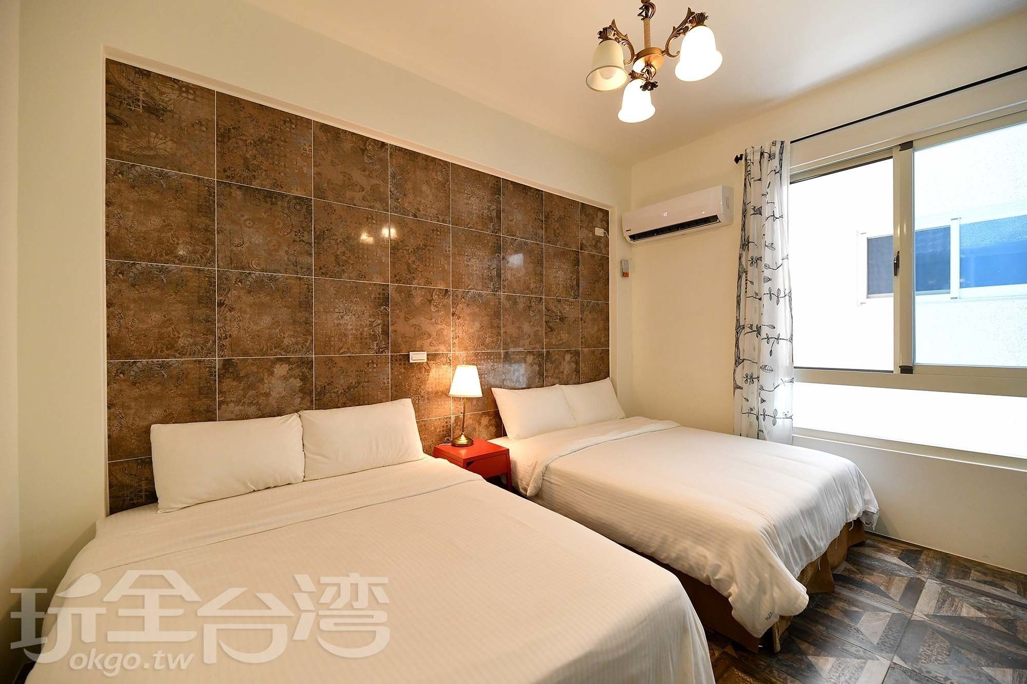 Une chambre d’hôtel avec deux lits, une grande fenêtre avec des rideaux, un lustre et un mur recouvert de carreaux décoratifs.