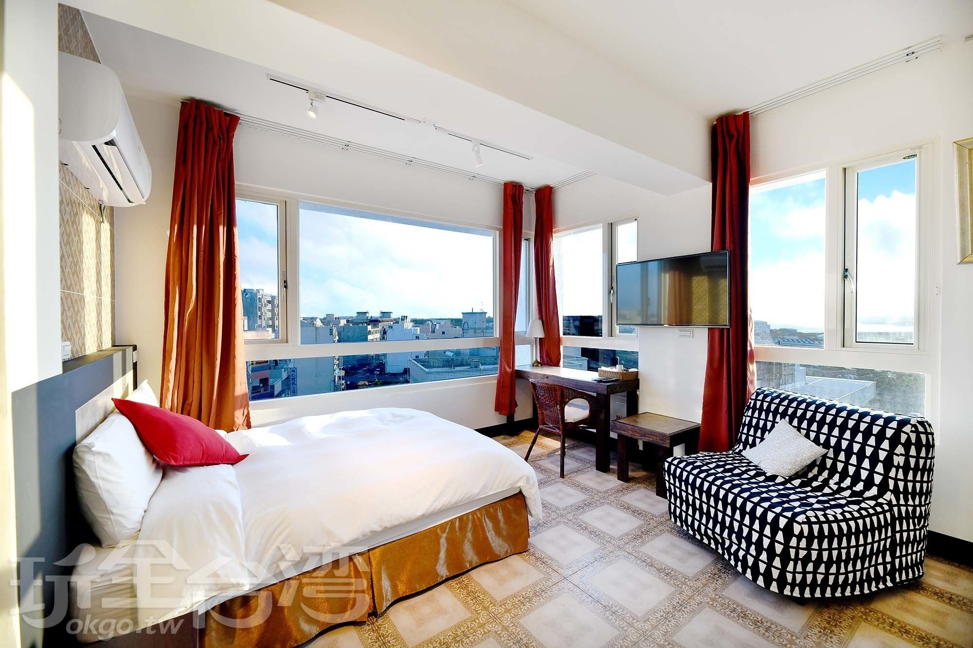 A hotel room with a bed, a desk, a chair, and a TV. There are large windows with curtains, and an air conditioning unit is mounted on the wall.