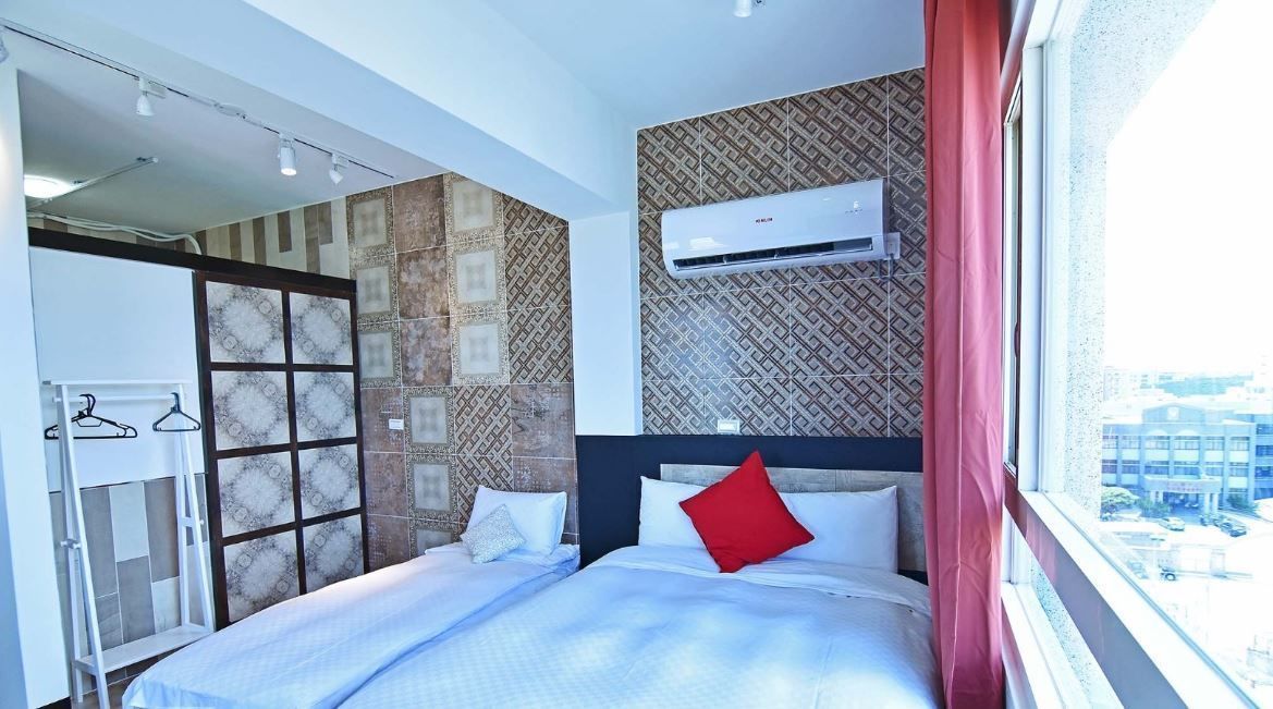 A hotel room with a bed, a wardrobe, an air conditioning unit, and a window with a city view.