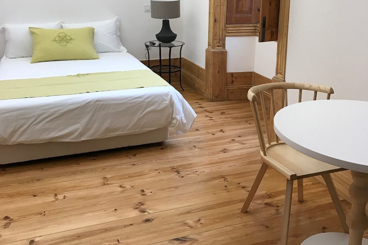 A room with a bed, a small table with a lamp, a wooden chair, and a wooden door.